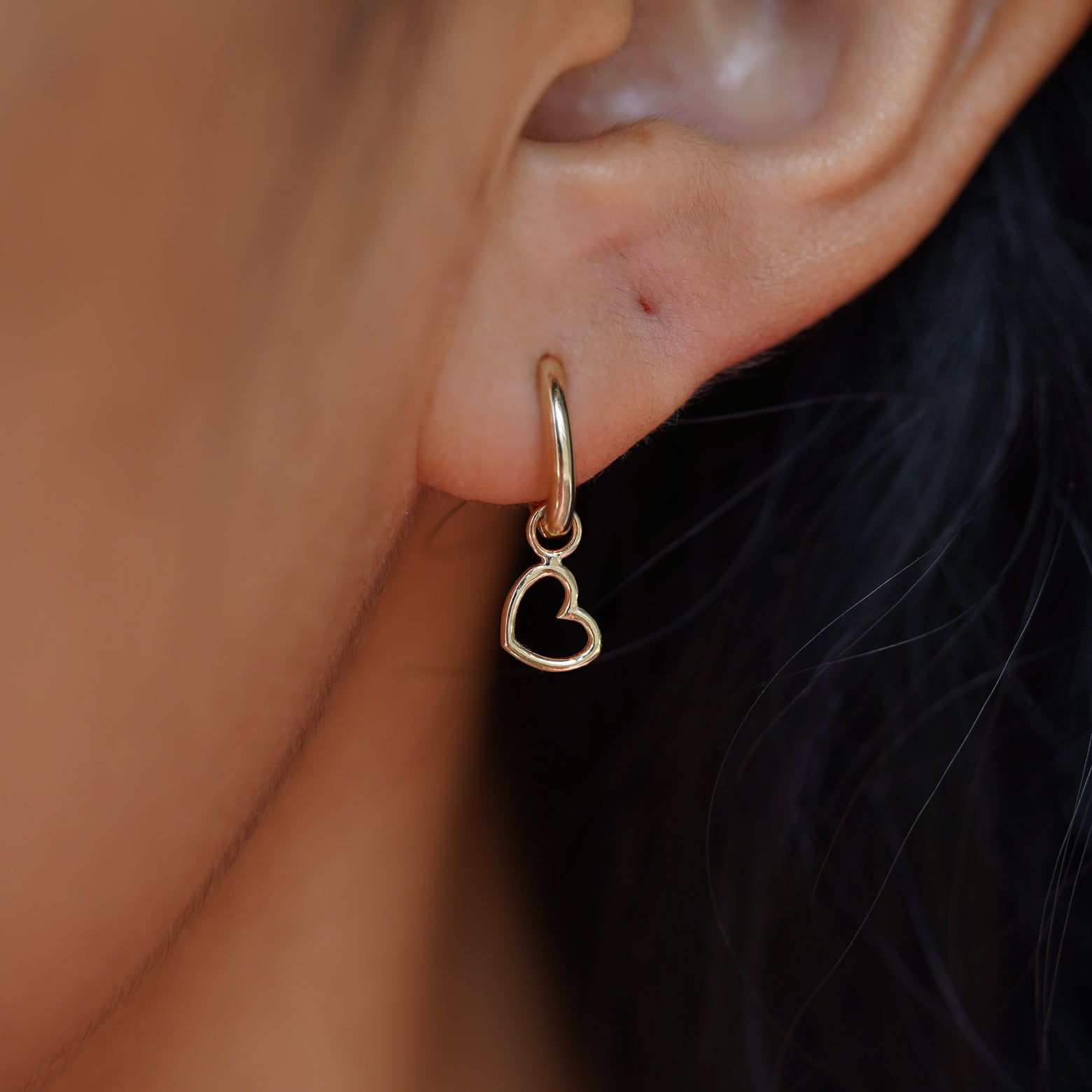 Close up view of a model's ear wearing a yellow gold Heart Charm on a Curvy Huggie Hoop