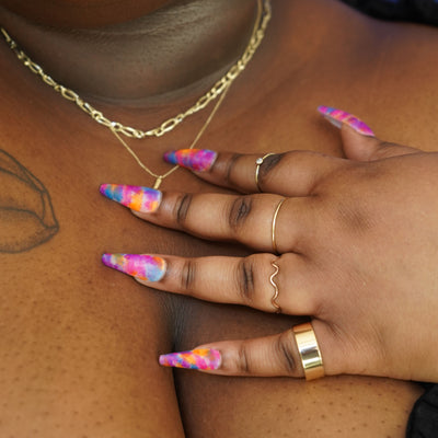 A model with long colorful nails touching an Automic Gold necklace around their neck while wearing Automic Gold rings
