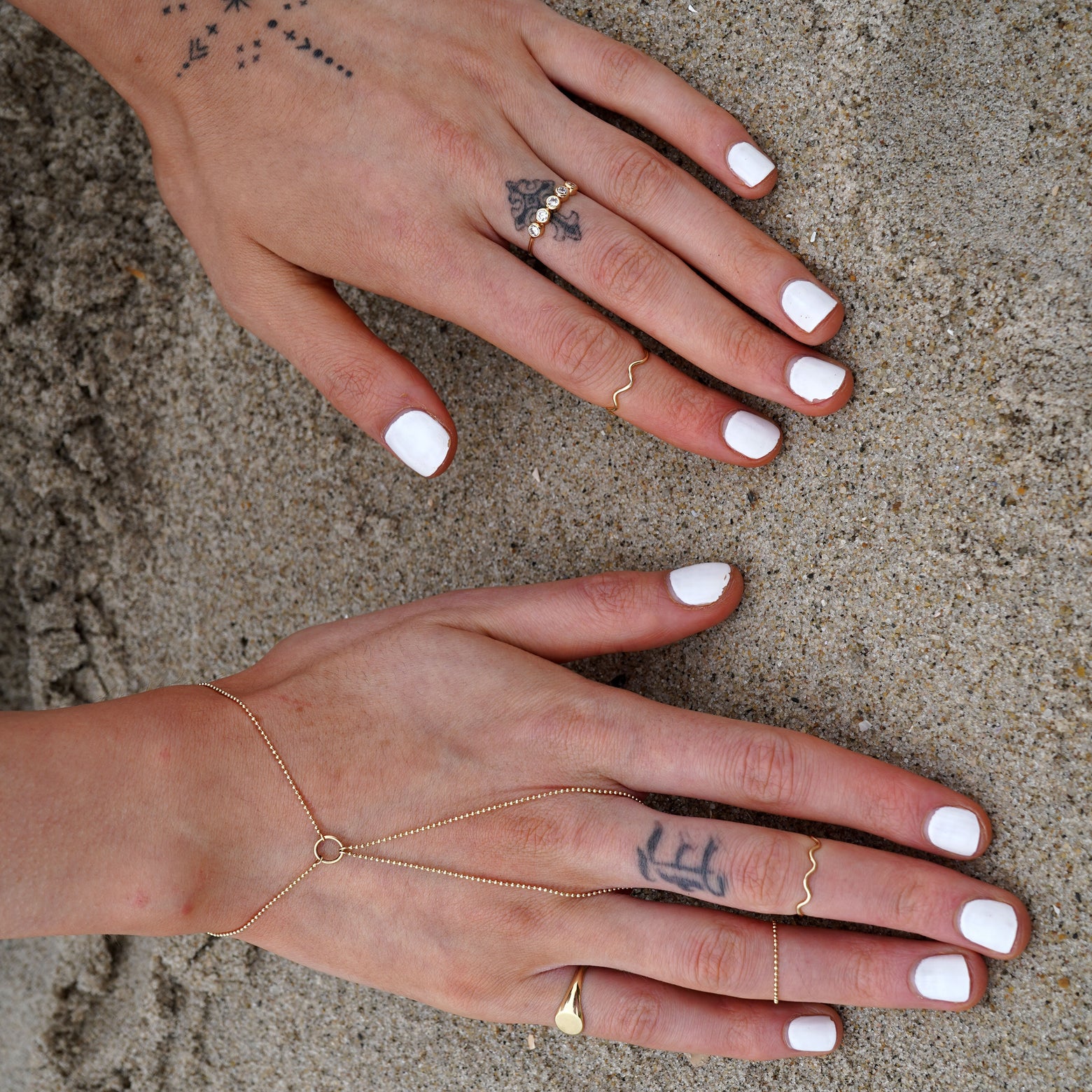A model with white nail polish on the beach with their hands in the sand while wearing Automic Gold rings and bracelets