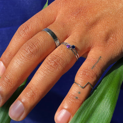 A model's tattooed hand wearing A Zig Zag ring, an Amethyst Ring, a Sapphire Ring, and an Industrial Wood Band in white gold